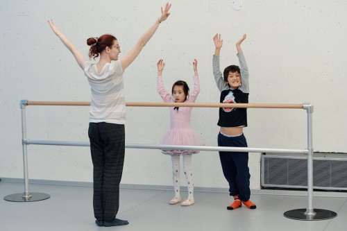 Two children, arms raised, participating to a NCDT's ballet class with their teacher in the studio
